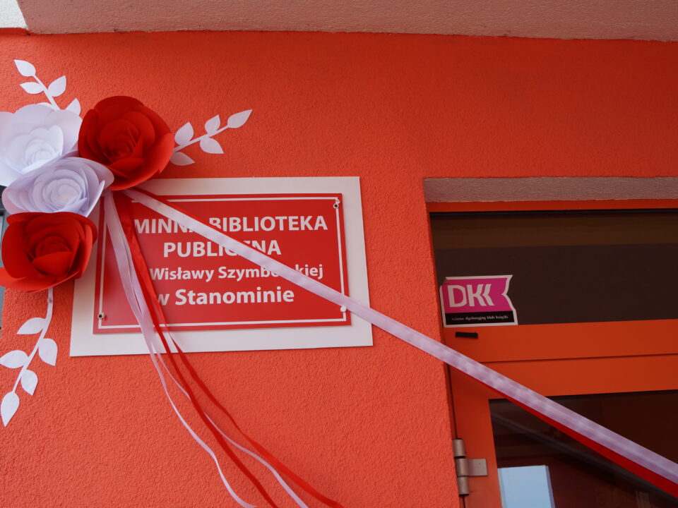 Tablica czerwona na białym tle na budynku biblioteki w Stanominie z napisem Gminna Biblioteka Publiczna im. Wisławy Szymborskiej w Stanominie